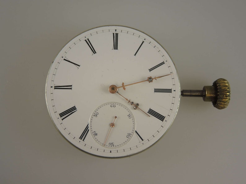 May include: A white pocket watch with Roman numeral hour markers and a smaller sub-dial for seconds. The watch has a gold-colored winding key and hands.