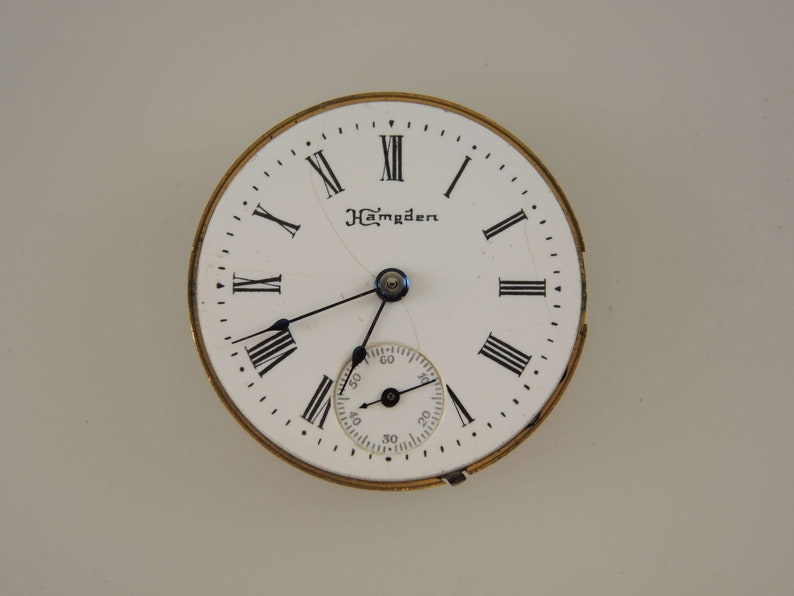 Puede incluir: Una esfera de reloj blanca con marcadores de hora en n&uacute;meros romanos negros y agujas negras. La esfera del reloj tiene la palabra "Hamden" impresa.