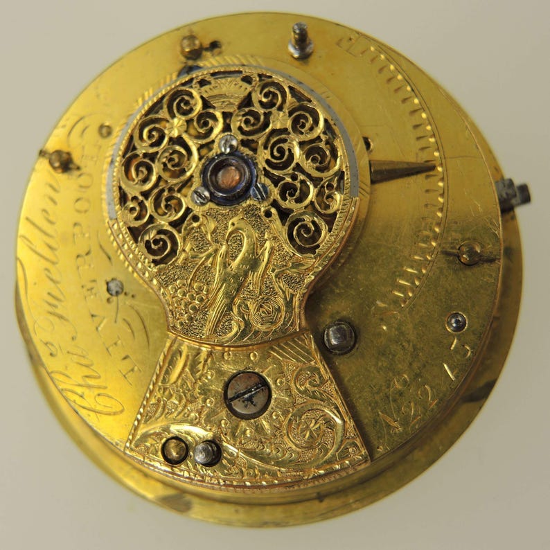 May include: Close-up of a vintage gold watch movement. The intricate mechanism features ornate filigree, engraved details, and visible gears. The watch face has a bird and floral design. The text "Cha. Welden" is visible.