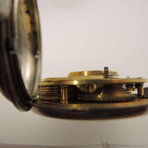 May include: Close-up of the inner workings of a vintage pocket watch. The watch's open case reveals intricate brass gears and mechanisms. The watch case is silver-toned, and the gears are a warm gold color.