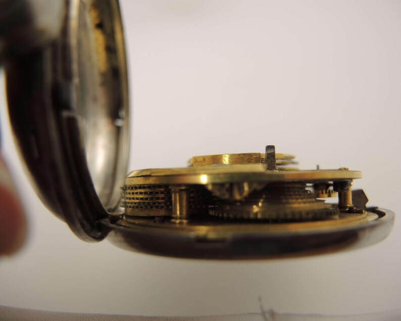May include: Close-up of the inner workings of a vintage pocket watch. The watch's open case reveals intricate brass gears and mechanisms. The watch case is silver-toned, and the gears are a warm gold color.