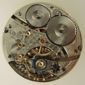 May include: Close-up of a vintage watch movement with intricate gears and components. The watch mechanism features the text "17 Jewels" and "A.W.W. Co. Waltham, Mass." The watch has a silver and gold color scheme.