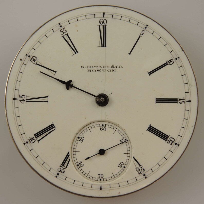 K&ouml;nnte beinhalten: Ein wei&szlig;es Taschenuhrenglas mit schwarzen r&ouml;mischen Ziffern f&uuml;r die Stunden und schwarzen Markierungen f&uuml;r die Minuten. Das Zifferblatt der Uhr hat einen kleineren, konzentrischen Kreis mit schwarzen Minutenmarkierungen und einem schwarzen Zeiger. Das Zifferblatt der Uhr tr&auml;gt au&szlig;erdem den Text "E. HOWARD & CO. BOSTON."