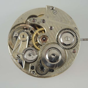 May include: Close-up of a silver pocket watch movement with exposed gears and a winding stem. The watch face is not visible.