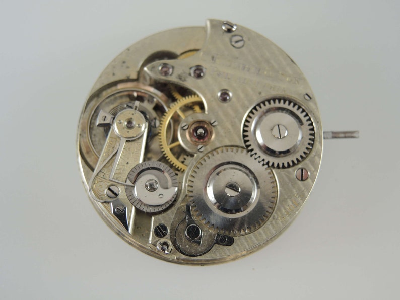 May include: Close-up of a silver pocket watch movement with exposed gears and a winding stem. The watch face is not visible.