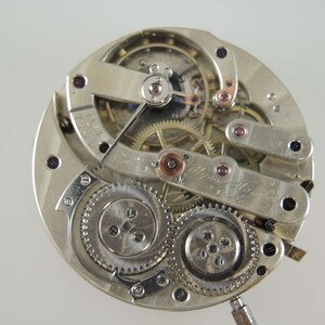 Op de afbeelding: Een close-up van een zilveren zakhorloge-uurwerk met ingewikkelde tandwielen en hendels. Het horloge is gedemonteerd en de interne mechanismen zijn zichtbaar. De wijzerplaat van het horloge is niet zichtbaar in deze afbeelding.