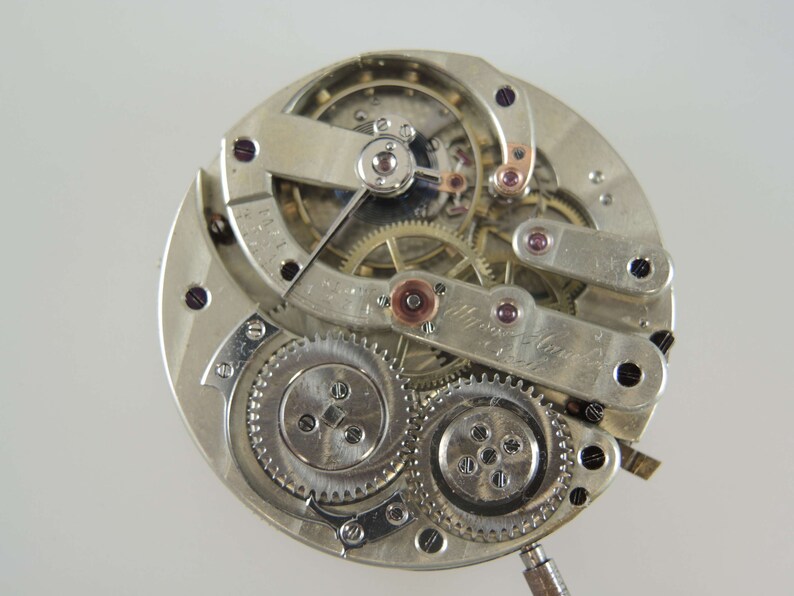 Op de afbeelding: Een close-up van een zilveren zakhorloge-uurwerk met ingewikkelde tandwielen en hendels. Het horloge is gedemonteerd en de interne mechanismen zijn zichtbaar. De wijzerplaat van het horloge is niet zichtbaar in deze afbeelding.