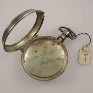 Könnte beinhalten: Eine silberne Taschenuhr mit geöffnetem Deckel, die das Innenleben der Uhr zeigt. Die Uhr hat ein kleines, rundes Zifferblatt und ein schlichtes Design.