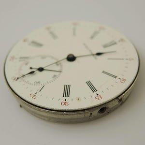 May include: Close-up of a vintage pocket watch face. The white face features black Roman numerals, red markers, and two smaller dials. The watch hands are black, and the metal casing is silver.