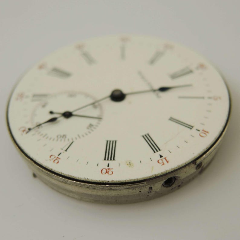May include: Close-up of a vintage pocket watch face. The white face features black Roman numerals, red markers, and two smaller dials. The watch hands are black, and the metal casing is silver.