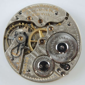May include: Close-up of a silver pocket watch movement with gold gears. The watch is labelled "Hamilton Watch Co. Lancaster Pa. 21 Jewels".