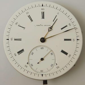May include: A white watch face with black Roman numerals and a smaller subdial. The watch face has the text "CHAS. JAGOT" and features black and gold hands. The outer edge is marked with small black lines indicating seconds.