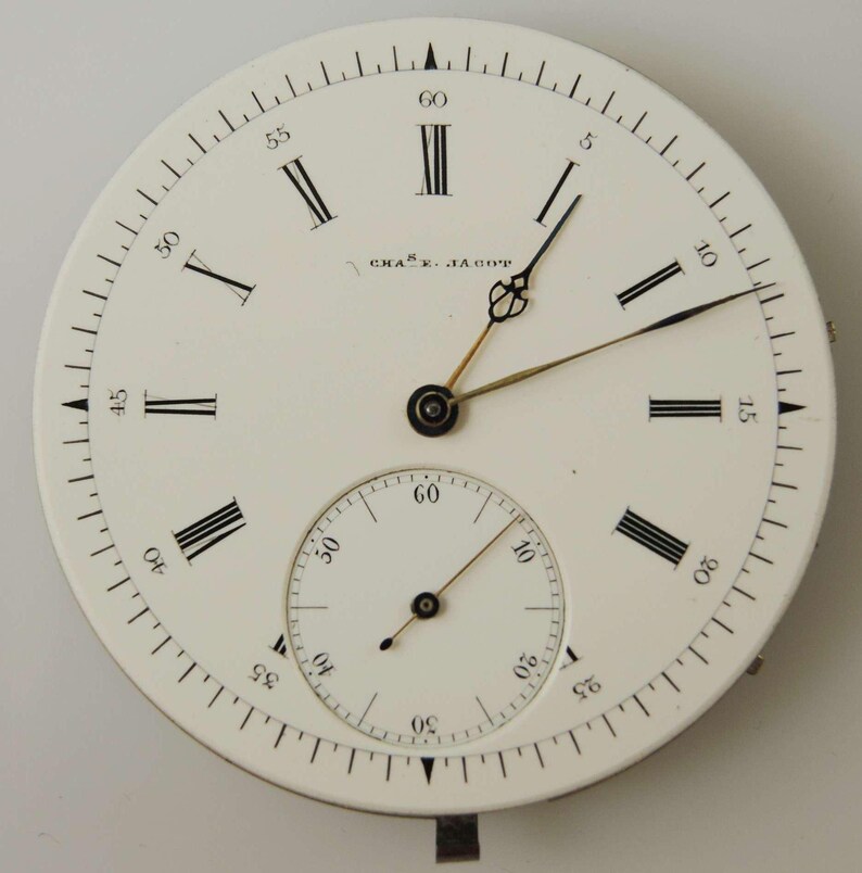 May include: A white watch face with black Roman numerals and a smaller subdial. The watch face has the text "CHAS. JAGOT" and features black and gold hands. The outer edge is marked with small black lines indicating seconds.