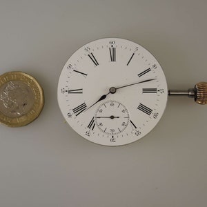 Puede incluir: Una esfera de reloj de bolsillo blanca con marcadores de hora en n&uacute;meros romanos negros y marcadores de minutos negros. El reloj tiene un subdial circular m&aacute;s peque&ntilde;o para los segundos. El reloj est&aacute; unido a un v&aacute;stago de cuerda con una perilla de color dorado.