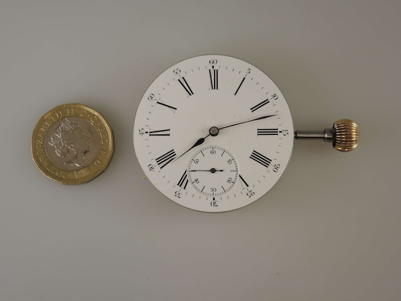 Puede incluir: Una esfera de reloj de bolsillo blanca con marcadores de hora en n&uacute;meros romanos negros y marcadores de minutos negros. El reloj tiene un subdial circular m&aacute;s peque&ntilde;o para los segundos. El reloj est&aacute; unido a un v&aacute;stago de cuerda con una perilla de color dorado.