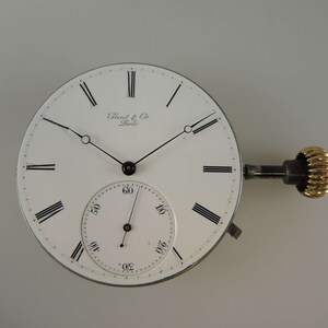 May include: A white pocket watch face with black Roman numeral hour markers and black hands. The watch face has the text "Dent & Co. London" printed on it. The watch is attached to a winding key with a gold-colored knob.