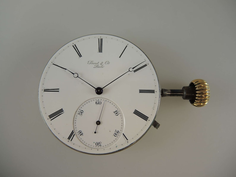 May include: A white pocket watch face with black Roman numeral hour markers and black hands. The watch face has the text "Dent & Co. London" printed on it. The watch is attached to a winding key with a gold-colored knob.