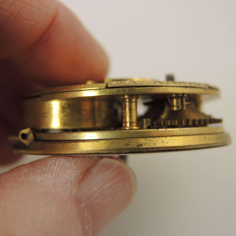 Puede incluir: Primer plano del mecanismo de un reloj de bolsillo antiguo. Se ven los componentes de lat&oacute;n, incluidos los engranajes y el volante. El reloj se muestra de lado, mostrando su intrincado funcionamiento interno y la p&aacute;tina envejecida.