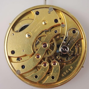 May include: Close-up of a gold-colored watch movement with intricate gears and a winding mechanism. The movement is labeled "Brevet&eacute; S.G.D.G." and "60648".