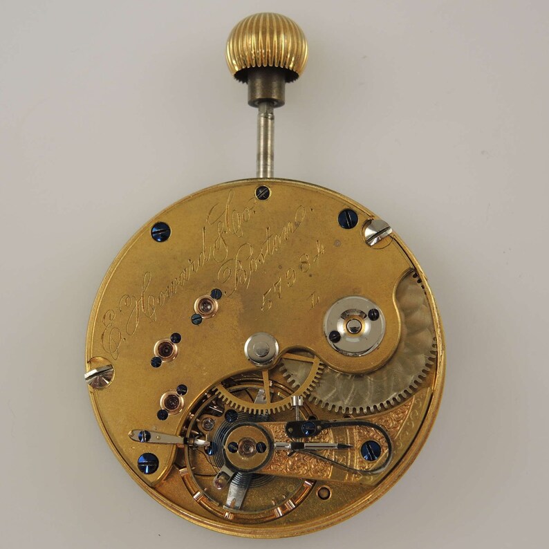 Puede incluir: Primer plano de un movimiento de reloj de bolsillo de oro con la inscripci&oacute;n "E. Howard & Co. Boston" y el n&uacute;mero "57454".