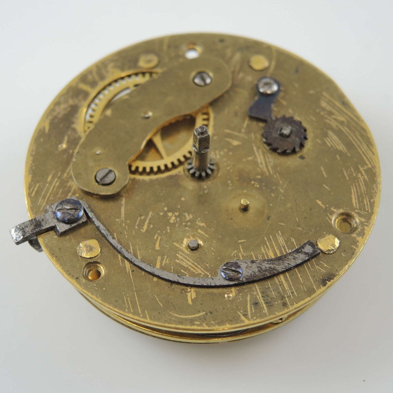 Op de afbeelding: Close-up van een goudkleurig uurwerk met ingewikkelde tandwielen en een zilverkleurige hendel. Het uurwerk is geplaatst tegen een effen witte achtergrond.