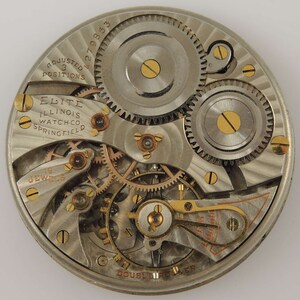 Op de afbeelding: Close-up van een zilveren zakhorloge-uurwerk met vergulde tandwielen en schroeven. Het horloge is gelabeld "Elite Illinois Watch Co. Springfield" en "Adjusted 3 Positions".