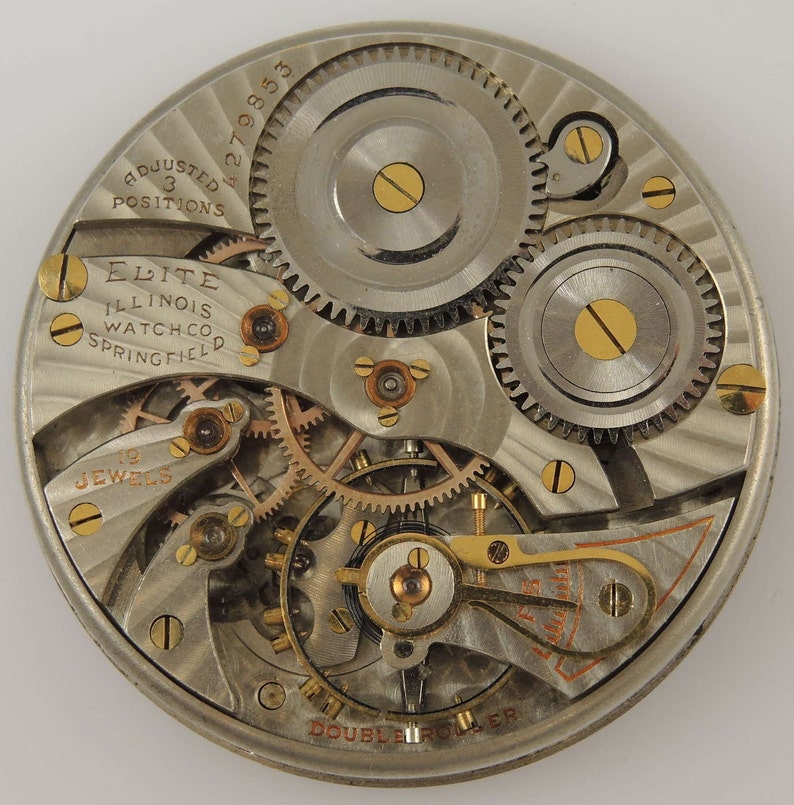 Op de afbeelding: Close-up van een zilveren zakhorloge-uurwerk met vergulde tandwielen en schroeven. Het horloge is gelabeld "Elite Illinois Watch Co. Springfield" en "Adjusted 3 Positions".