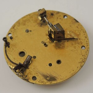 May include: Close-up of a gold-colored circular clock face with a small metal lever and a gear mechanism. The clock face has several holes and a small inscription.