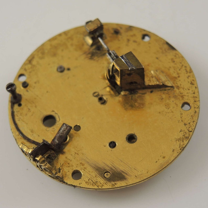 May include: Close-up of a gold-colored circular clock face with a small metal lever and a gear mechanism. The clock face has several holes and a small inscription.