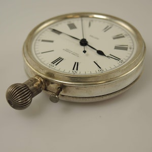 K&ouml;nnte beinhalten: Eine silberfarbene Taschenuhr mit wei&szlig;em Zifferblatt und schwarzen r&ouml;mischen Ziffern. Die Uhr hat einen strukturierten Aufzugsknopf und einen kleinen Einstellstift. Die Zeiger sind schwarz, vor schlichtem Hintergrund.