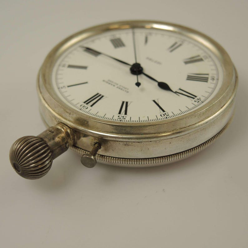 K&ouml;nnte beinhalten: Eine silberfarbene Taschenuhr mit wei&szlig;em Zifferblatt und schwarzen r&ouml;mischen Ziffern. Die Uhr hat einen strukturierten Aufzugsknopf und einen kleinen Einstellstift. Die Zeiger sind schwarz, vor schlichtem Hintergrund.