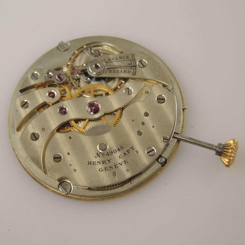 May include: A detailed view of a vintage watch movement, showcasing intricate gears, jewels, and screws. The mechanism is silver with gold-coloured gears and a gold crown. The text "AVANCE RETARD" is visible, along with "No 49048 HENRY CAPT GENEVE".