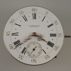 May include: A white pocket watch face with black Roman numeral hour markers and black minute markers. The watch face has a gold-colored rim and hands. The watch face also has the text "S.R. SMITH SON PHILADELPHIA." printed on it.