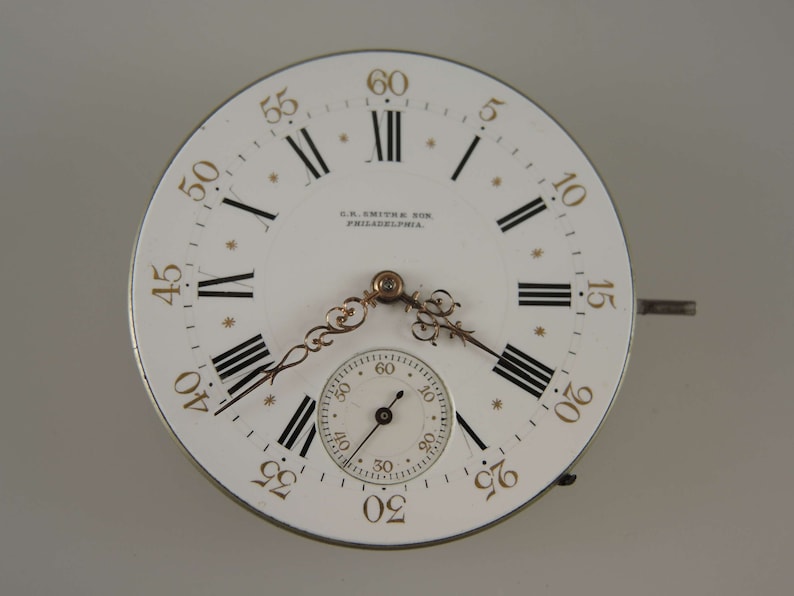 May include: A white pocket watch face with black Roman numeral hour markers and black minute markers. The watch face has a gold-colored rim and hands. The watch face also has the text "S.R. SMITH SON PHILADELPHIA." printed on it.