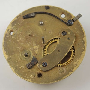 May include: Close-up of a brass clock mechanism with a gold gear and a curved metal piece. The mechanism is mounted on a round brass plate.