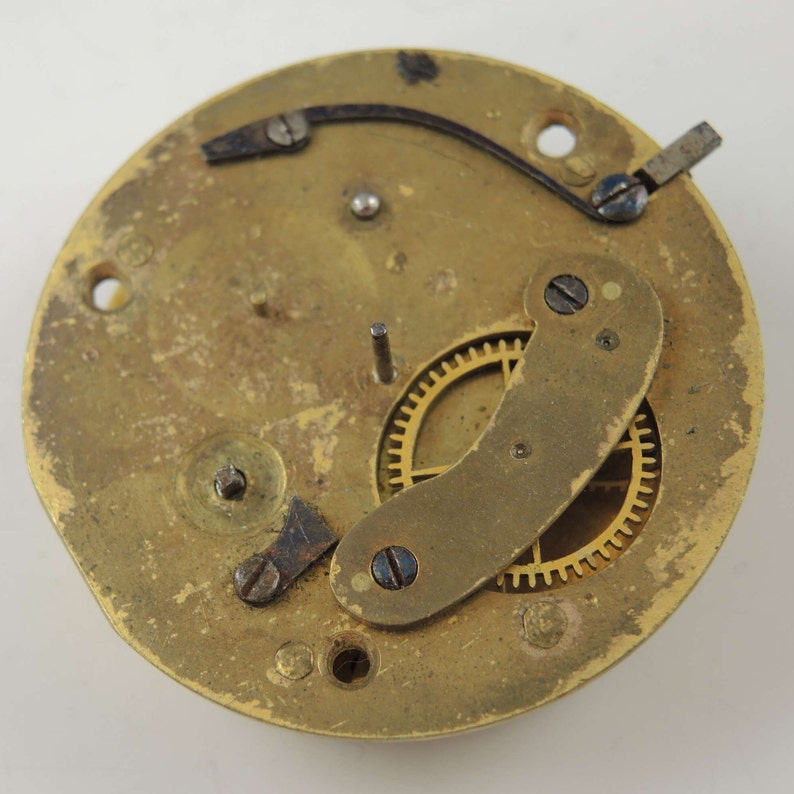 May include: Close-up of a brass clock mechanism with a gold gear and a curved metal piece. The mechanism is mounted on a round brass plate.