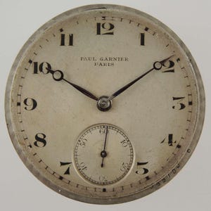 May include: A vintage watch face with a cream-colored dial and black numerals. The watch face has the text "PAUL GARNIER PARIS" and a small seconds dial. The hands are black, and the watch has a silver-colored metal casing.