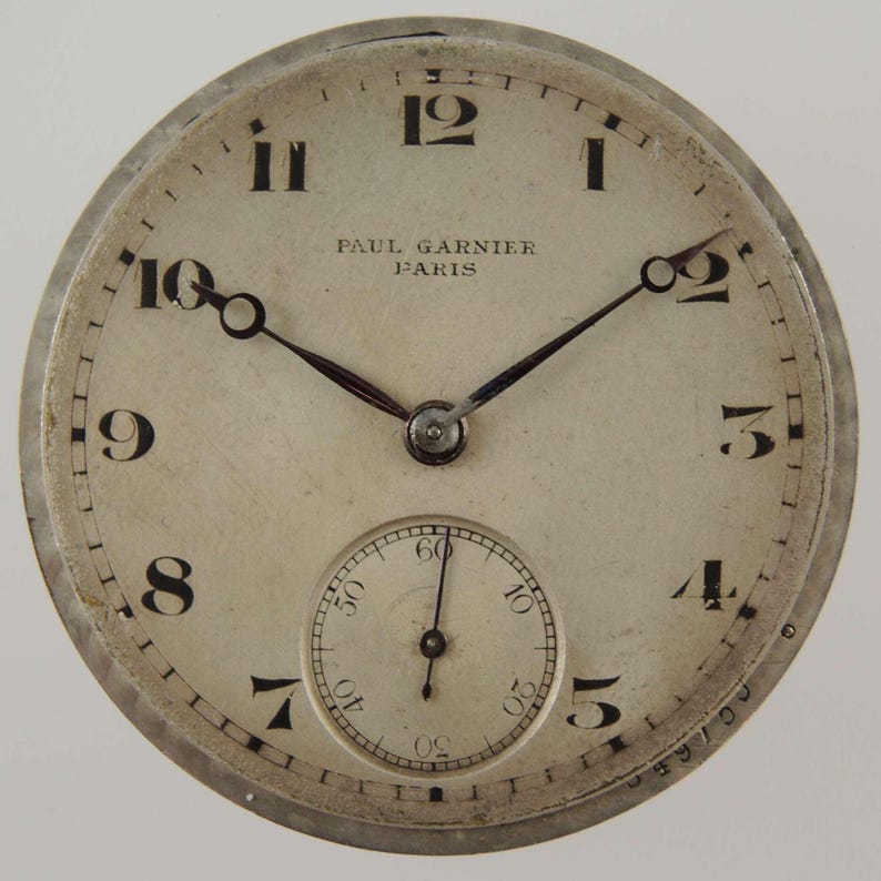 May include: A vintage watch face with a cream-colored dial and black numerals. The watch face has the text "PAUL GARNIER PARIS" and a small seconds dial. The hands are black, and the watch has a silver-colored metal casing.