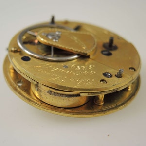 Puede incluir: Un movimiento de reloj de bolsillo de oro con una tapa parcialmente abierta. El movimiento est&aacute; inscrito con el texto "J.C. & S.  London  No. 1120".