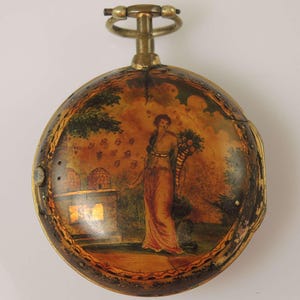 May include: Vintage pocket watch with a hand-painted scene. The watch face depicts a woman in a long dress holding flowers, set in a garden with beehives. The watch has a gold-coloured case and a winding stem.