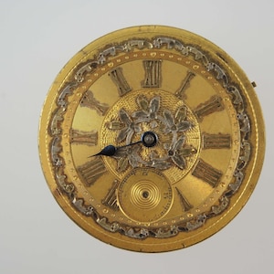 May include: Close-up of a gold-toned pocket watch face with Roman numerals and ornate detailing. The watch face features a smaller subdial and delicate floral accents. The hands are black, and the overall design is antique.
