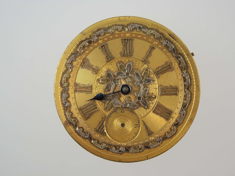 May include: Close-up of a gold-toned pocket watch face with Roman numerals and ornate detailing. The watch face features a smaller subdial and delicate floral accents. The hands are black, and the overall design is antique.