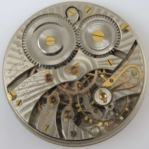 Op de afbeelding: Close-up van de binnenkant van een zilveren zakhorloge. Het horloge is gelabeld met "Elite Illinois Watch Co. Springfield" en "Adjusted 3 Positions 19853".