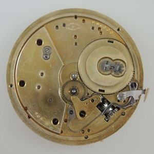 May include: Close-up view of the inner workings of a gold-toned pocket watch. The watch face is removed, revealing intricate gears and a winding mechanism.