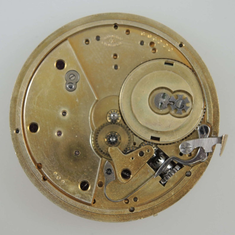 May include: Close-up view of the inner workings of a gold-toned pocket watch. The watch face is removed, revealing intricate gears and a winding mechanism.