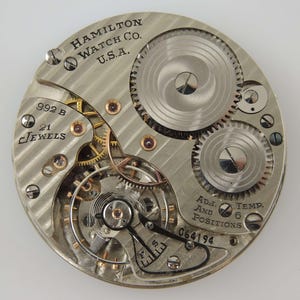 May include: Detailed view of a Hamilton Watch Co. U.S.A. watch movement. The silver mechanism displays intricate gears, jewels, and the text "992 B" and "21 Jewels". Additional text includes "Adj. And Positions" and "Temp. 6".