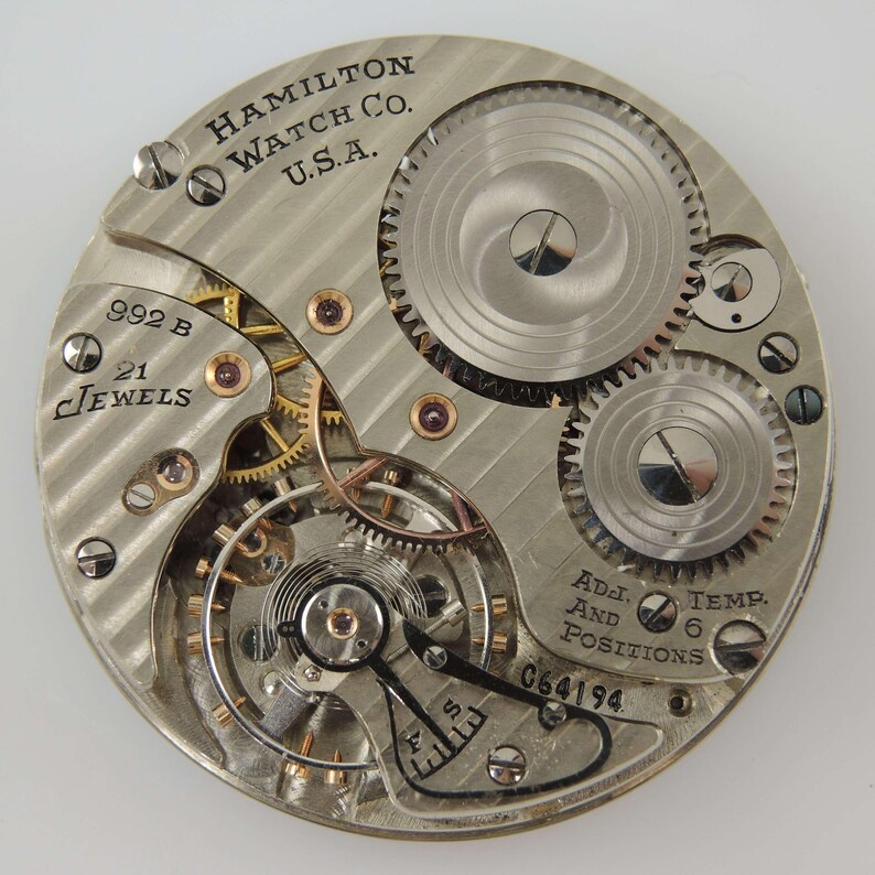 May include: Detailed view of a Hamilton Watch Co. U.S.A. watch movement. The silver mechanism displays intricate gears, jewels, and the text "992 B" and "21 Jewels". Additional text includes "Adj. And Positions" and "Temp. 6".