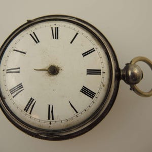 May include: A vintage pocket watch with a white face and black Roman numerals. The watch has a silver-colored casing and a gold-colored ring for attaching a chain. The hands are black and the watch shows the time as approximately 2:40.