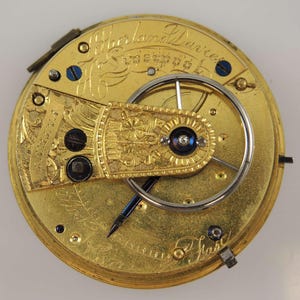 May include: Close-up of a gold pocket watch with intricate engravings. The watch face is visible, showing the inner workings of the mechanism. The inscription "Sherland Davies Liverpool" is visible on the watch face.
