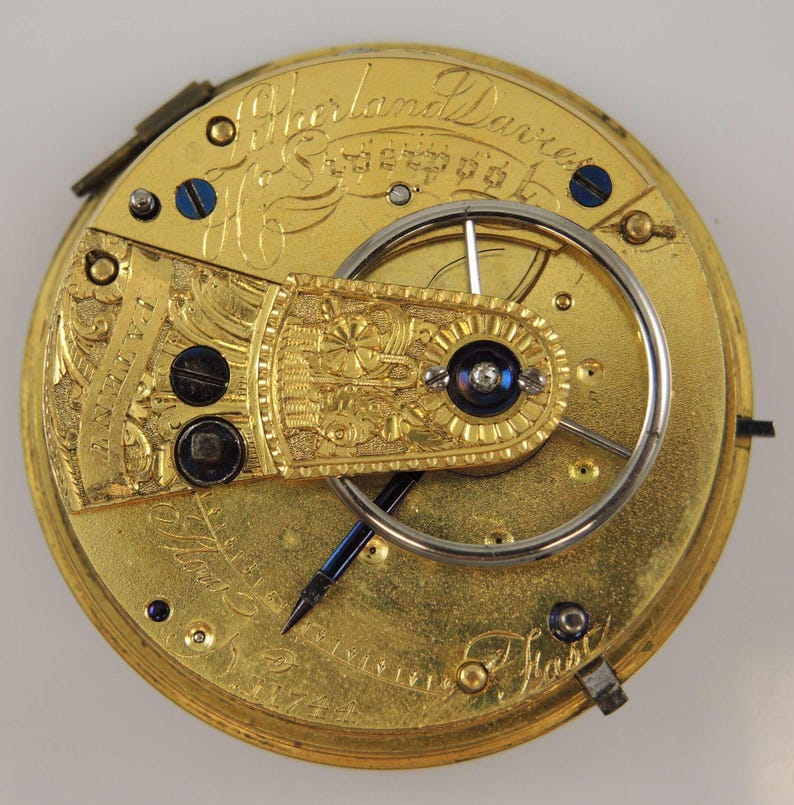 May include: Close-up of a gold pocket watch with intricate engravings. The watch face is visible, showing the inner workings of the mechanism. The inscription "Sherland Davies Liverpool" is visible on the watch face.